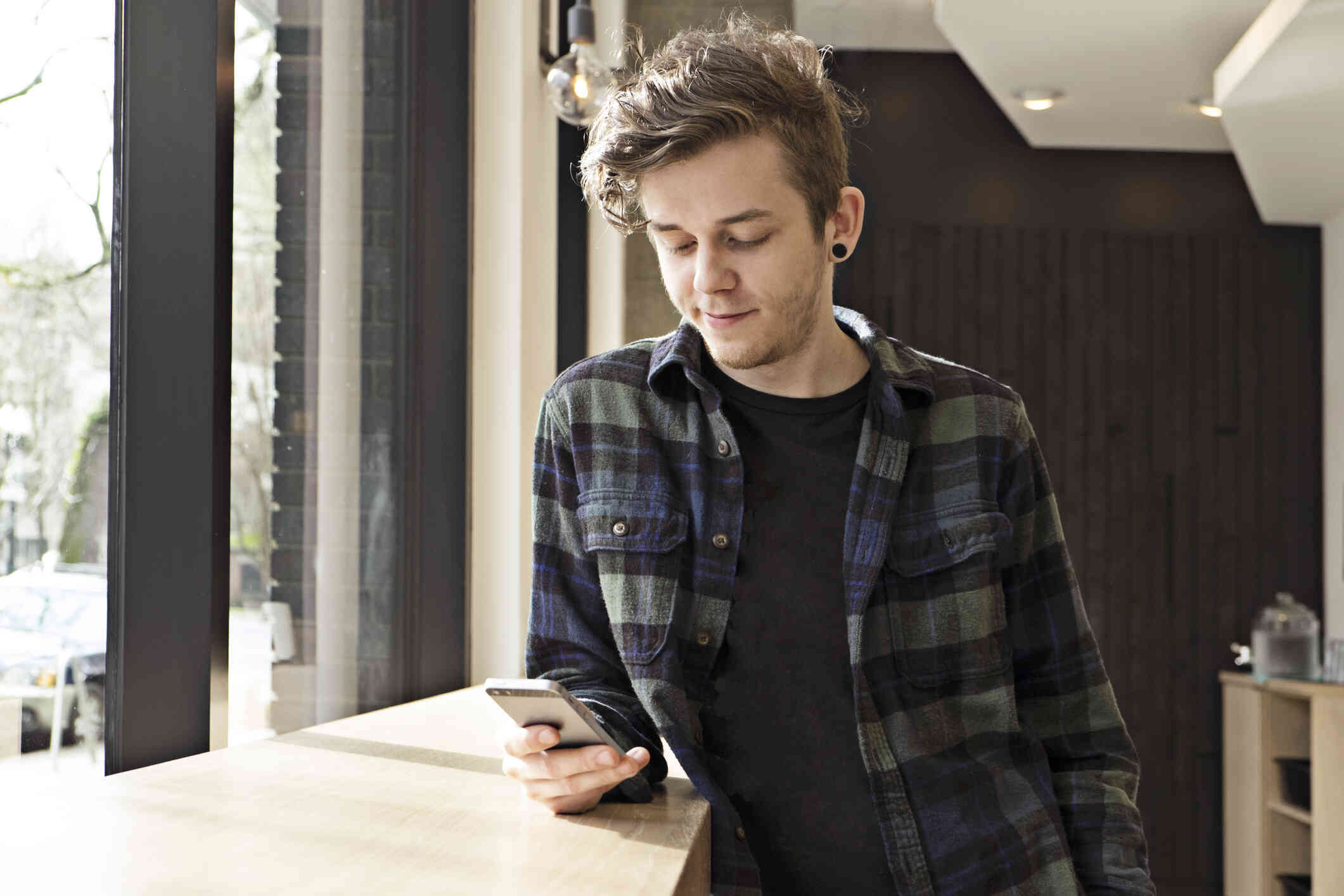 A man in a plaid shirt leans against a counter near a wndow and looks down at the cellphone in his hand.